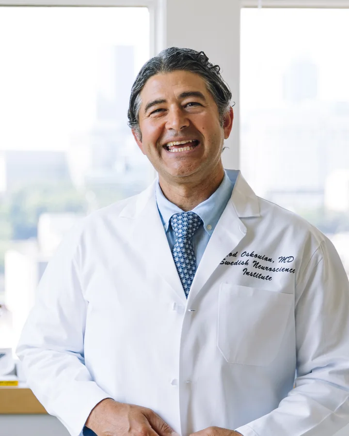 Rod Oskouian laughing in a white coat embroidered with his name and Swedish Neuroscience Institute, by a bright office window